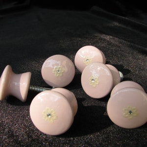 May include: Six pink ceramic drawer knobs with a white floral design and a single rhinestone in the center of each flower.