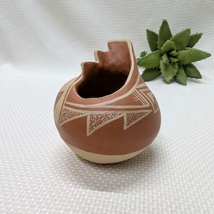 May include: A handcrafted, terracotta-colored pottery vessel with a unique, stepped rim. The round body features geometric designs in a lighter, cream color. The vessel is likely used for decorative purposes.