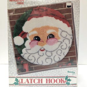 May include: A WonderArt by Caron International latch hook kit box featuring a circular Santa Claus design. The graphic shows a round Santa face with a red hat, white beard, and pink cheeks against a brick background. The box is labeled Santa 27 inches.