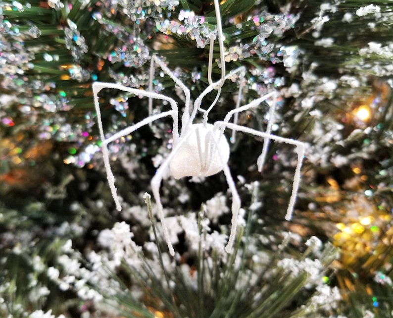 May include: A white, glitter-covered spider Christmas ornament with long, spindly legs. The spider is hanging from a Christmas tree with green needles and white, snowy accents. The background has colorful, blurred lights.