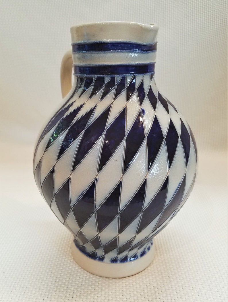 May include: A white ceramic pitcher with a blue and white diamond pattern. The pitcher has a handle and a wide mouth.