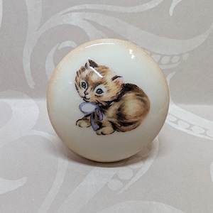 May include: A round, cream-colored cabinet knob featuring a charming illustration of a kitten with brown and tan fur, wearing a purple bow. The knob has a glossy finish and is suitable for furniture.