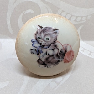 May include: A round, off-white ceramic knob featuring a charming illustration of a gray and white kitten wearing a blue bow. The kitten is seated next to a pink ball of yarn. The knob has a glossy finish.