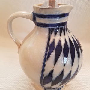 May include: A white ceramic pitcher with a blue and white diamond pattern. The pitcher has a handle and a spout.