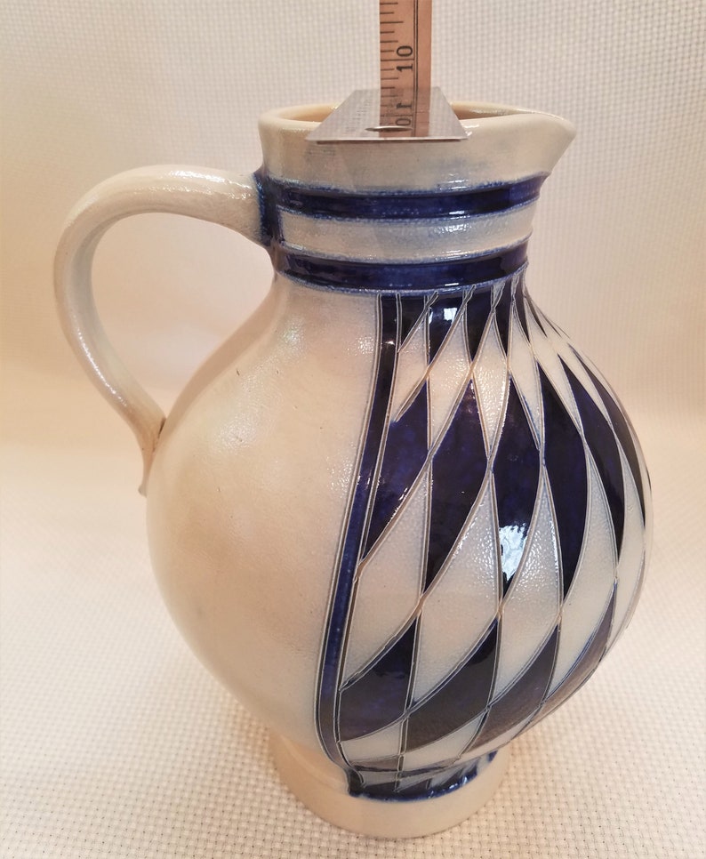 May include: A white ceramic pitcher with a blue and white diamond pattern. The pitcher has a handle and a spout.