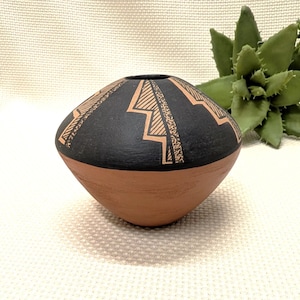 May include: A handcrafted, small, round pottery vase. The vase has a terracotta-colored base and a black upper portion with geometric, tan-colored designs. The vase is a decorative item, suitable for displaying small flowers or as a standalone art piece.