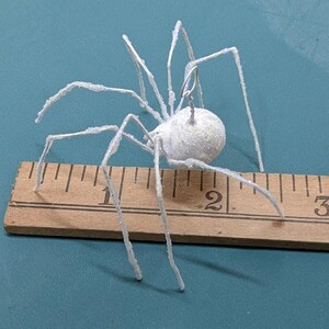 May include: A white, glitter-covered spider decoration with wire legs, measuring approximately 2 inches in length. The spider is positioned on a wooden ruler against a teal background, suitable for Halloween or themed decor.