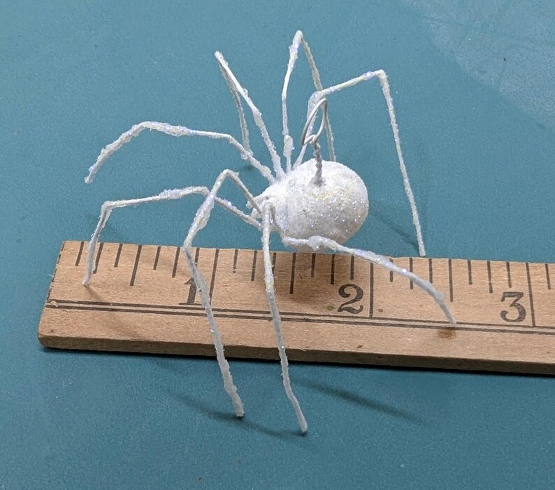 May include: A white, glitter-covered spider decoration with wire legs, measuring approximately 2 inches in length. The spider is positioned on a wooden ruler against a teal background, suitable for Halloween or themed decor.