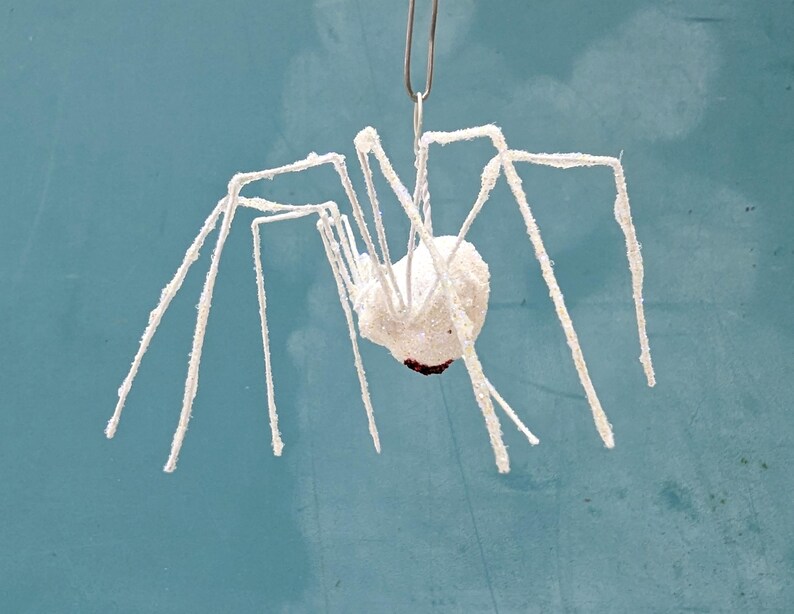 May include: A white, glitter-covered spider decoration with long, slender legs. The spider is suspended by a silver clip against a teal background. The spider's body is a round shape with a dark brown spot.