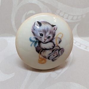 May include: A round, off-white cabinet knob featuring a charming illustration of a gray and white kitten with a blue bow and a ball of yellow yarn. The knob has a glossy finish and is likely made of ceramic.