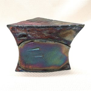May include: A ceramic box with a geometric shape and a iridescent finish. The box has a lid that fits snugly on top. The box is made of a dark gray clay with a metallic sheen.
