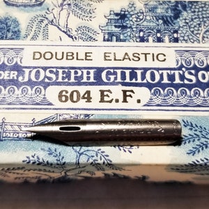 May include: A silver-colored double elastic pen nib with the text "Joseph Gillott's Own Selection" and "604 E.F." on the box. The box has a blue and white floral pattern.