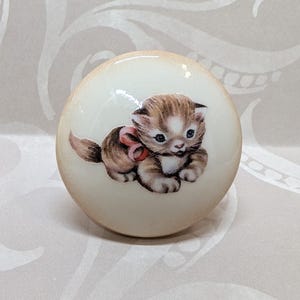 May include: A round, off-white cabinet knob featuring a detailed illustration of a brown and white kitten with a pink bow. The knob has a glossy finish and a light brown edge, suitable for furniture or decor.
