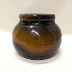 May include: A small, round, brown ceramic pot with a dark brown glaze. The pot has a slightly rounded lip and a smooth, glossy finish.