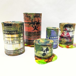 May include: Five rusty metal canisters with biohazard symbols and warning labels. The canisters are green, red, blue, and brown with black text. The canisters are sitting on a white surface.