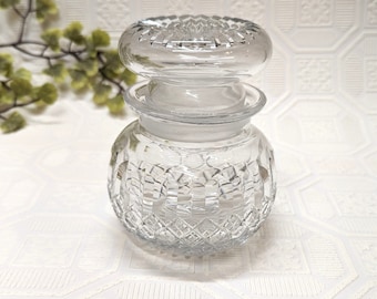 May include: A clear glass jar with a textured, diamond-shaped pattern. The jar has a lid with a raised, circular design.