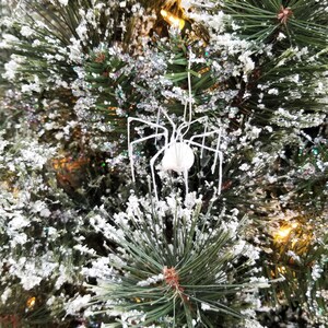 May include: A white spider ornament with long, thin legs hangs from a snow-covered Christmas tree. The tree's green needles are dusted with white snow and glitter, creating a festive holiday scene. The spider has a round body.
