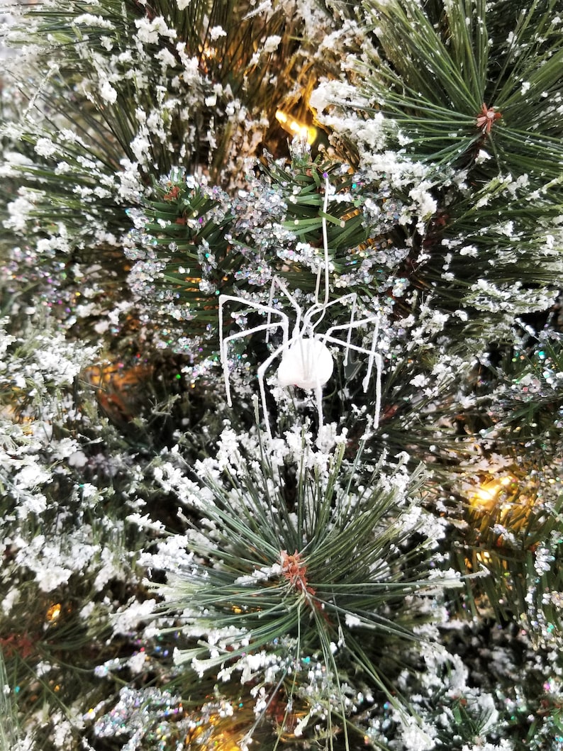 May include: A white spider ornament with long, thin legs hangs from a snow-covered Christmas tree. The tree's green needles are dusted with white snow and glitter, creating a festive holiday scene. The spider has a round body.