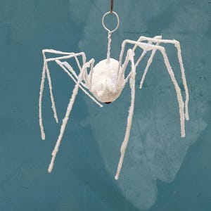 May include: A white, glitter-covered spider ornament with long, spindly legs. The spider is suspended by a silver wire loop, perfect for hanging. The spider's body is a round shape with a red accent. Ideal for Halloween decorations.