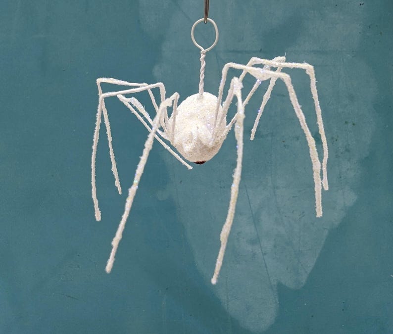 May include: A white, glitter-covered spider ornament with long, spindly legs. The spider is suspended by a silver wire loop, perfect for hanging. The spider's body is a round shape with a red accent. Ideal for Halloween decorations.