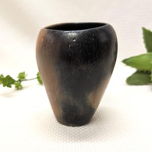 May include: A handcrafted, dark-colored ceramic vase with a unique, asymmetrical shape. The vase features a blend of black and brown tones, creating a rustic and earthy aesthetic. The opening is oval-shaped. The vase is suitable for displaying flowers or as a decorative piece.