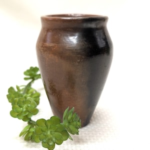 May include: A small, handcrafted brown vase with a dark, shaded finish. The vase has a rounded body and a slightly flared rim. A green succulent plant is placed near the vase.