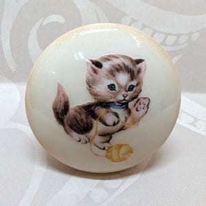 May include: A round, off-white knob featuring a charming illustration of a kitten playing with a ball of yarn. The kitten is depicted in shades of brown, gray, and white, with a blue collar. The knob has a slightly aged, vintage appearance.