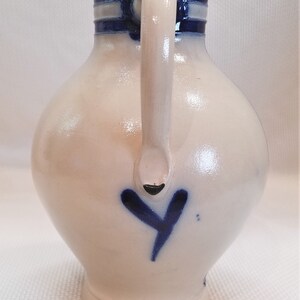 May include: A white ceramic pitcher with a blue and white striped handle and a blue heart design. The pitcher has a round body and a wide base.