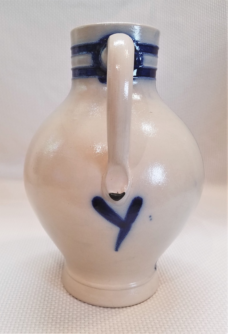 May include: A white ceramic pitcher with a blue and white striped handle and a blue heart design. The pitcher has a round body and a wide base.
