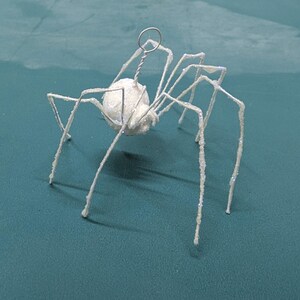 May include: A white, glitter-covered spider ornament with a round body and long, thin legs. The spider has a wire loop at the top for hanging. The ornament is set against a teal background.
