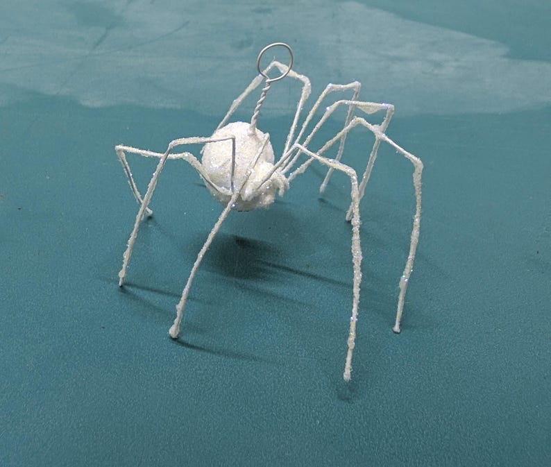 May include: A white, glitter-covered spider ornament with a round body and long, thin legs. The spider has a wire loop at the top for hanging. The ornament is set against a teal background.