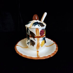 May include: A white ceramic honey pot with a lid and a small spoon. The pot is decorated with a floral design in shades of green, yellow, red, and brown.