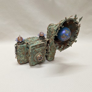 May include: A vintage-style camera made of metal with a green patina finish. The camera has intricate floral details and a decorative lens cover. A blue and gold orb is visible inside the lens cover.