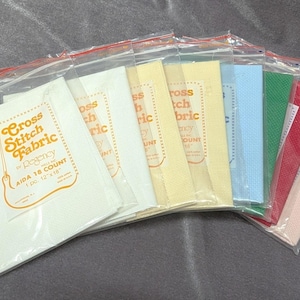 May include: A collection of cross-stitch fabric in clear plastic bags with red zip closures. The fabric is available in various colors, including white, yellow, blue, green, red, and pink. Each bag has a label with the text "Cross Stitch Fabric".