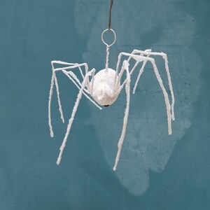 May include: A white, glitter-covered spider ornament with long, spindly legs. The spider is suspended by a thin wire and ring, against a teal background. The spider's body is a round, white ball with a small red accent.