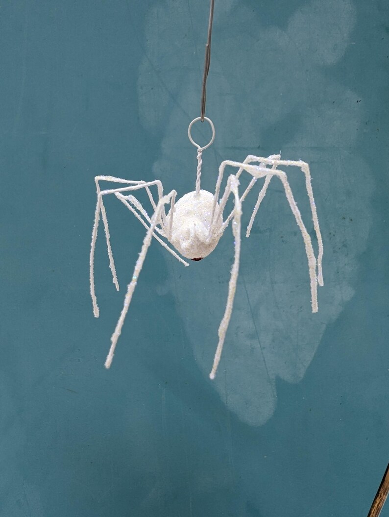 May include: A white, glitter-covered spider ornament with long, spindly legs. The spider is suspended by a thin wire and ring, against a teal background. The spider's body is a round, white ball with a small red accent.