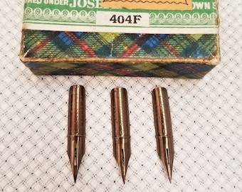 May include: Three vintage metal pen nibs with a box. The nibs are bronze-colored and have a pointed tip. The box has a green and plaid design with the text "SHEFFIELD. 3" and "404F".