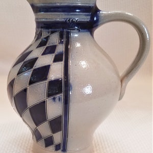 May include: A blue and white ceramic pitcher with a checkered pattern. The pitcher has a handle and a spout. It is a traditional German style.