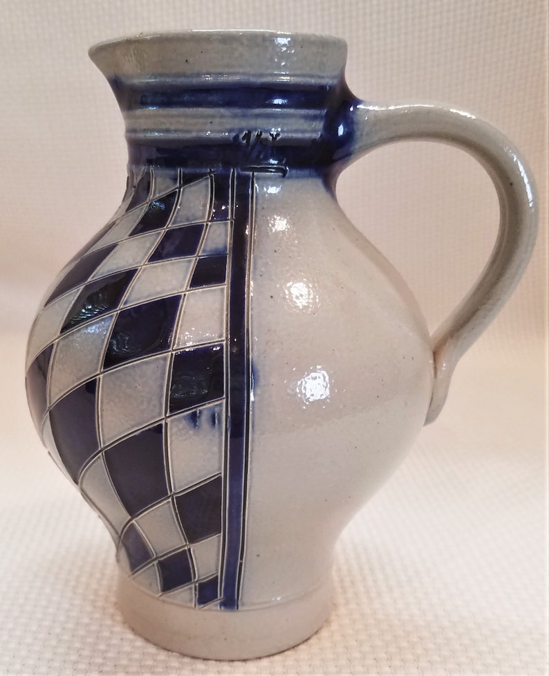 May include: A blue and white ceramic pitcher with a checkered pattern. The pitcher has a handle and a spout. It is a traditional German style.