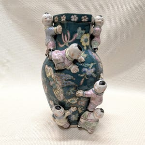 May include: A teal ceramic vase with a floral and butterfly design, adorned with six small, climbing figures in pastel clothing. The vase has a hexagonal top and is decorated with flowers.