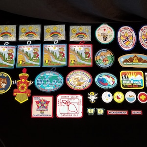 May include: A collection of colorful embroidered patches featuring various scout themes, including "Be Prepared", "Tiger Cub", "Camp Morrison", "Catalina Isle", and "Trail's End".