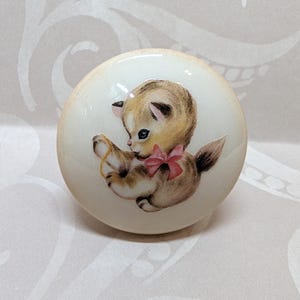 May include: A round, off-white cabinet knob featuring a vintage illustration of a kitten. The kitten is depicted in shades of brown and tan, with a pink bow. The knob is likely made of ceramic or porcelain.