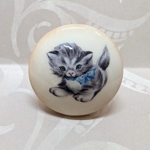 May include: A round, off-white cabinet knob featuring a gray and white kitten with a blue bow. The kitten is depicted in a vintage, illustrative style. The knob has a glossy finish and is suitable for furniture or decor.