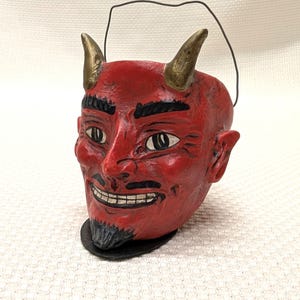 May include: A red devil head figurine with gold horns, black eyebrows, and a goatee. The devil has a sinister smile and a wire handle. The figurine is on a black base and is likely a Halloween decoration.