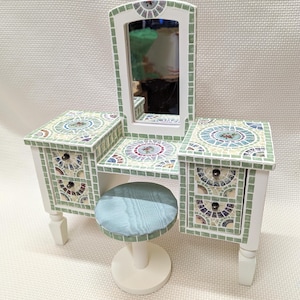 May include: A white vanity set with a mirror and two drawers on each side. The vanity top and drawers are covered in a mosaic of green, blue, and white tiles. A white stool with a light blue cushion sits in front of the vanity.