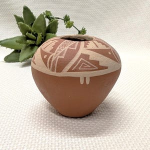 May include: A small, round, terracotta-colored pottery vase with a cream-colored geometric design around the top. The vase has a small opening and is displayed with a green succulent plant in the background.