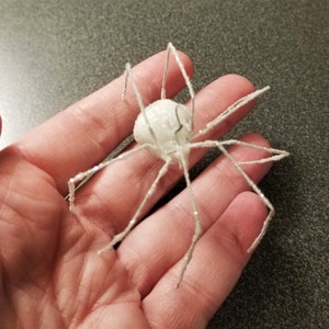 May include: A white, glittery spider decoration with long, silver legs and a round body. The spider is a Halloween or holiday-themed craft item, perfect for adding a spooky touch to any decor.