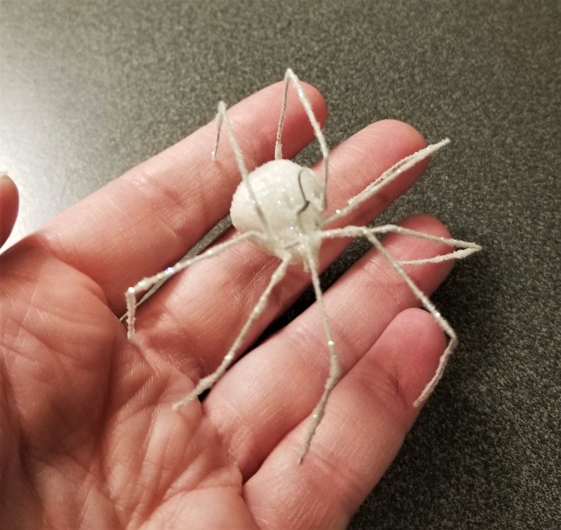May include: A white, glittery spider decoration with long, silver legs and a round body. The spider is a Halloween or holiday-themed craft item, perfect for adding a spooky touch to any decor.