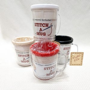 May include: Several clear glass mugs with white, beige, red, and black rims. Each mug has a beige fabric wrap and the words "STITCH A MUG" printed on a white label. The mugs are designed for cross-stitch projects.