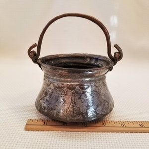 May include: A small, antique copper pot with a handle. The pot is about 4 inches tall and has a diameter of about 3 inches. The pot has a hammered finish and is decorated with a floral design.